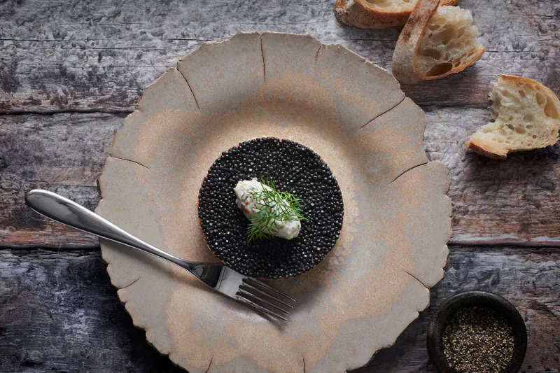 Close-up of caviar on plate with crème fraîche, herbs, bread chunks, fork, and pepper grains on wooden table