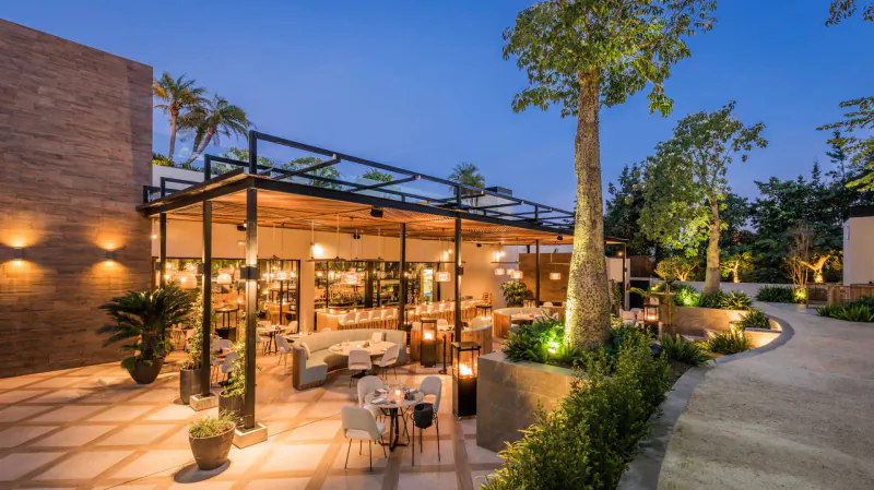 Twilight outdoor patio at Breathe Marbella Gallery with lounge seating, palm trees, lanterns, and lush landscaping.