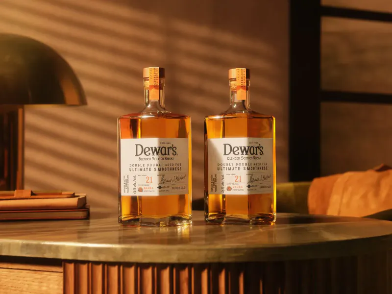 Two bottles of Dewar's Ultimate 21 whisky on a brass table with lamp, warm lighting.