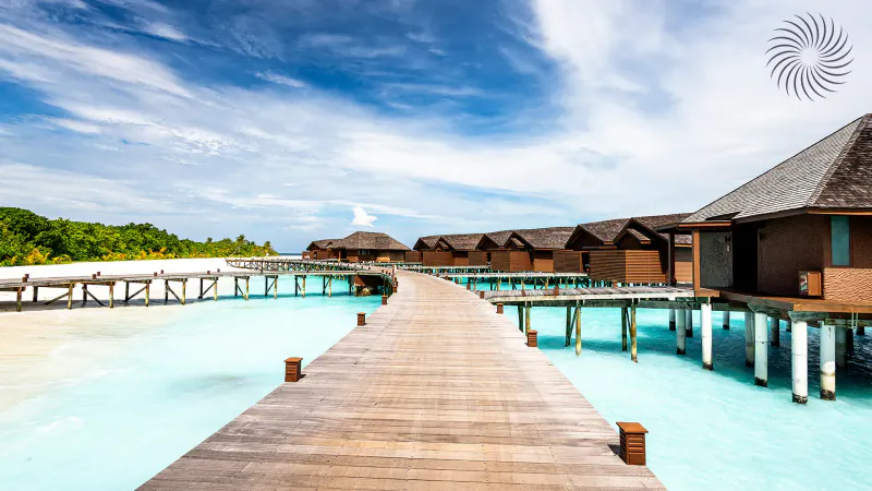 Wooden pier leading to overwater bungalows at Hideaway Beach Resort & Spa Maldives, turquoise lagoon, white sand, blue sky.