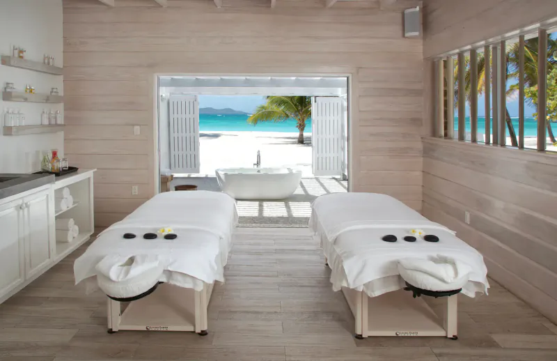 Two massage tables with towels and stones in spa room opening to white sand beach and turquoise sea at Palm Island Resort.