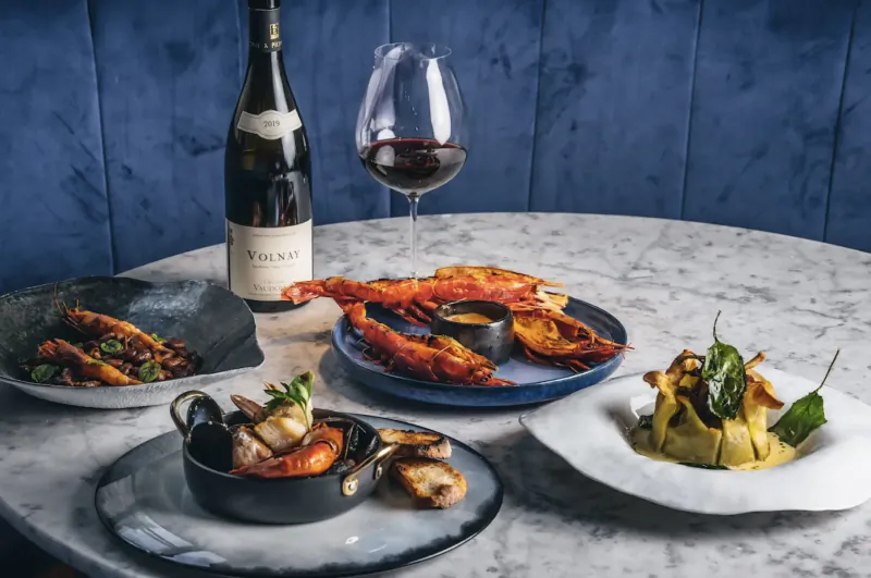 Elegant table at Cavo with red wine bottle and glass, lobster, mussels, and Mediterranean dishes on marble surface against blue velvet backdrop.