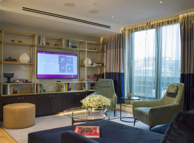 Elegant Guardsman hotel lounge with gold shelves, large purple TV, green armchairs, white flowers, and city view.