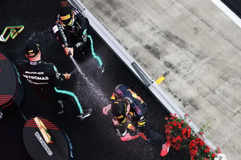 Overhead view of Mercedes mechanics in black suits hosing down a Red Bull driver in pink suit on F1 pit lane.