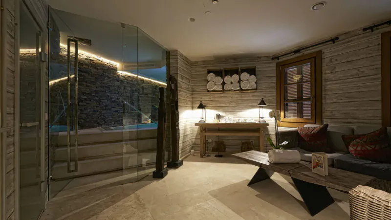 Cozy lounge in El Lodge Sierra Nevada with glass-walled indoor pool, rustic decor, lamps, sofa, and coffee table.