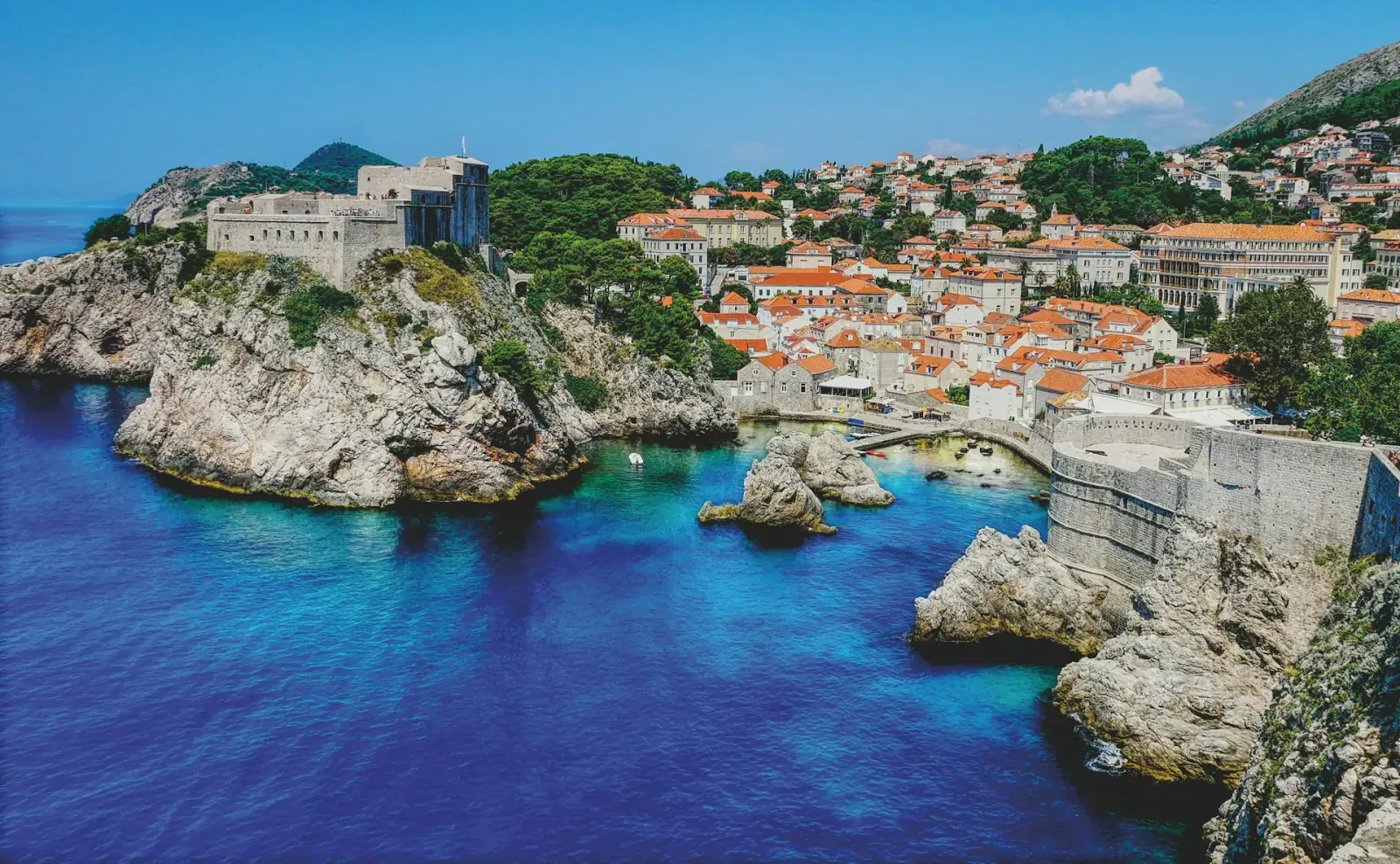 Aerial view of Dubrovnik's old town with red-roofed buildings, stone walls, cliffs, and turquoise Adriatic Sea.