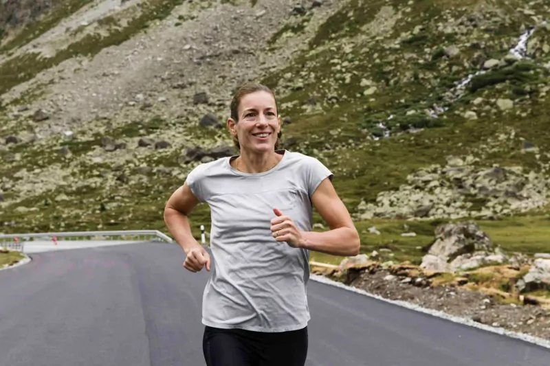 Female triathlete Nicola smiling while running on mountain road with rocky peaks