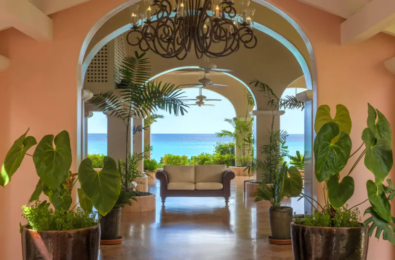 Pink arched hotel corridor with chandelier, potted palms, sofa, and ocean view at Fairmont Royal Pavilion.