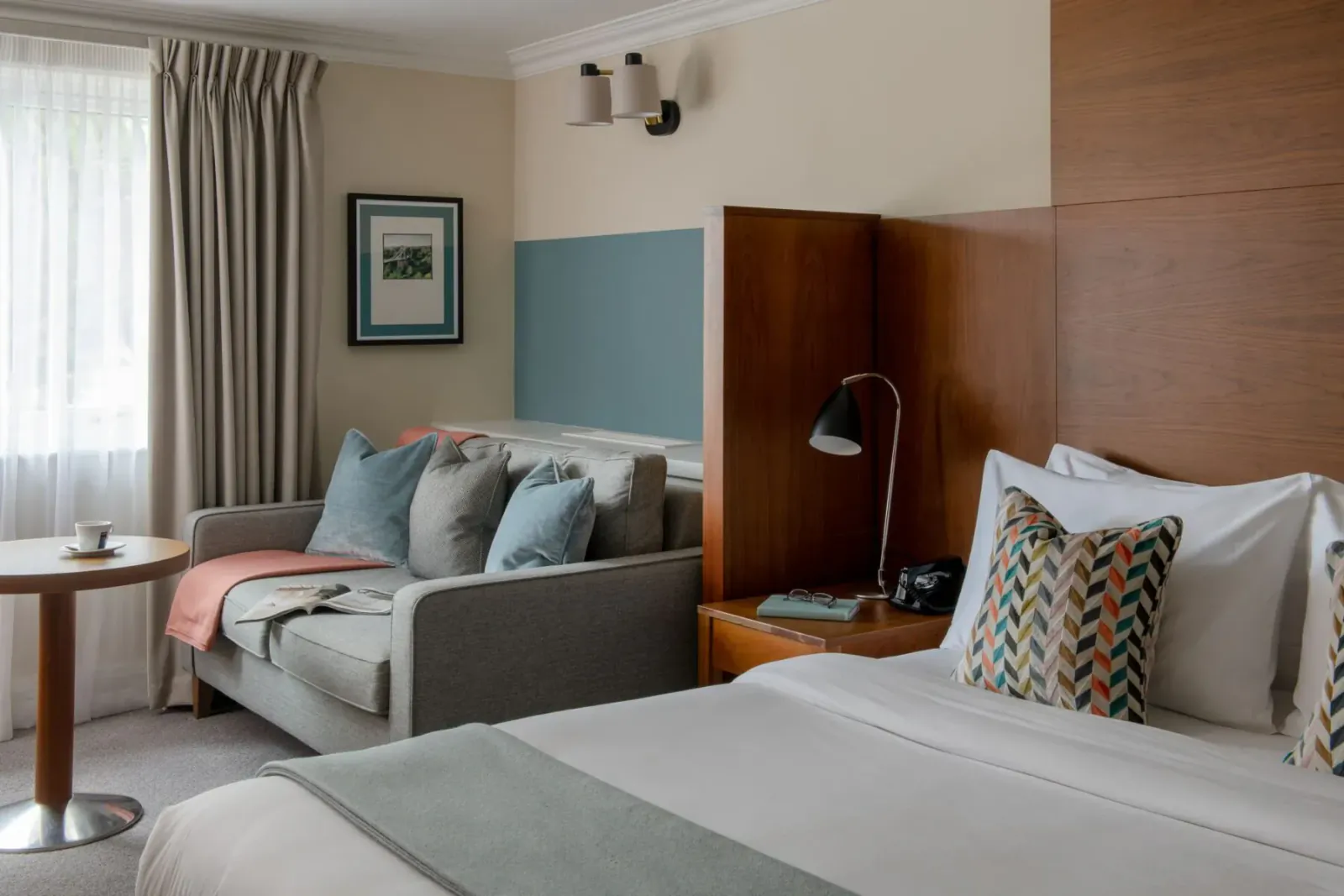 Cozy hotel room with plush bed, colorful pillows, gray sofa, lamp, and window drapes.
