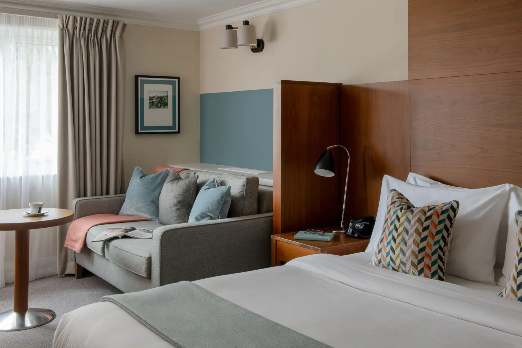 Cozy hotel room with plush bed, colorful pillows, gray sofa, lamp, and window drapes.