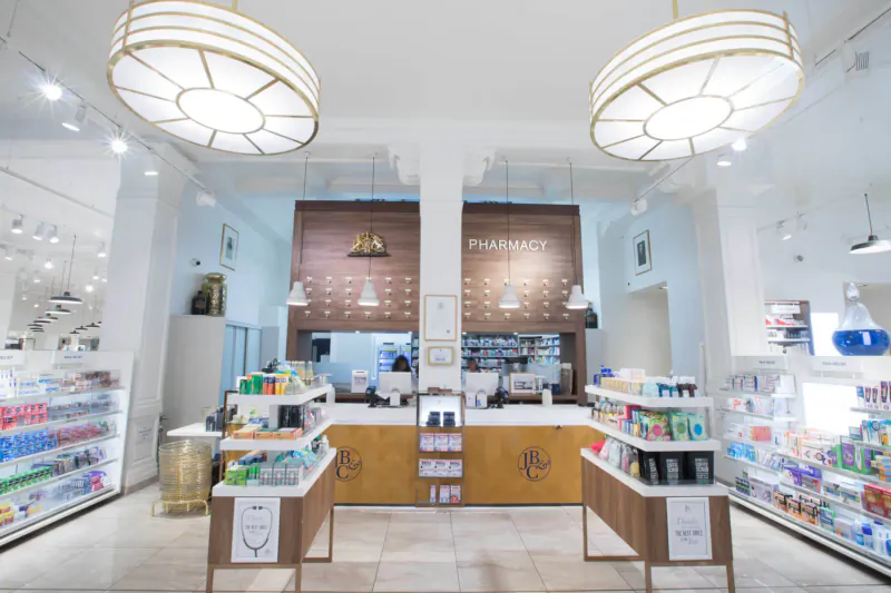 Spacious modern pharmacy interior in Marylebone Village with wooden counter, shelves of products, and gold pendant lights.