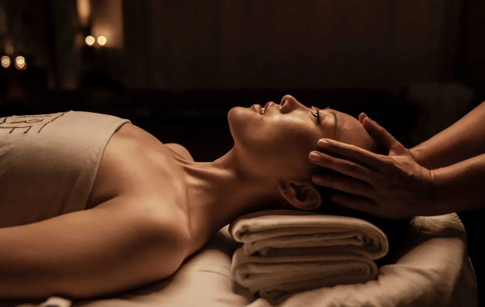 Woman lying on spa bed at Aire Ancient Baths, receiving relaxing head massage in candlelit room.