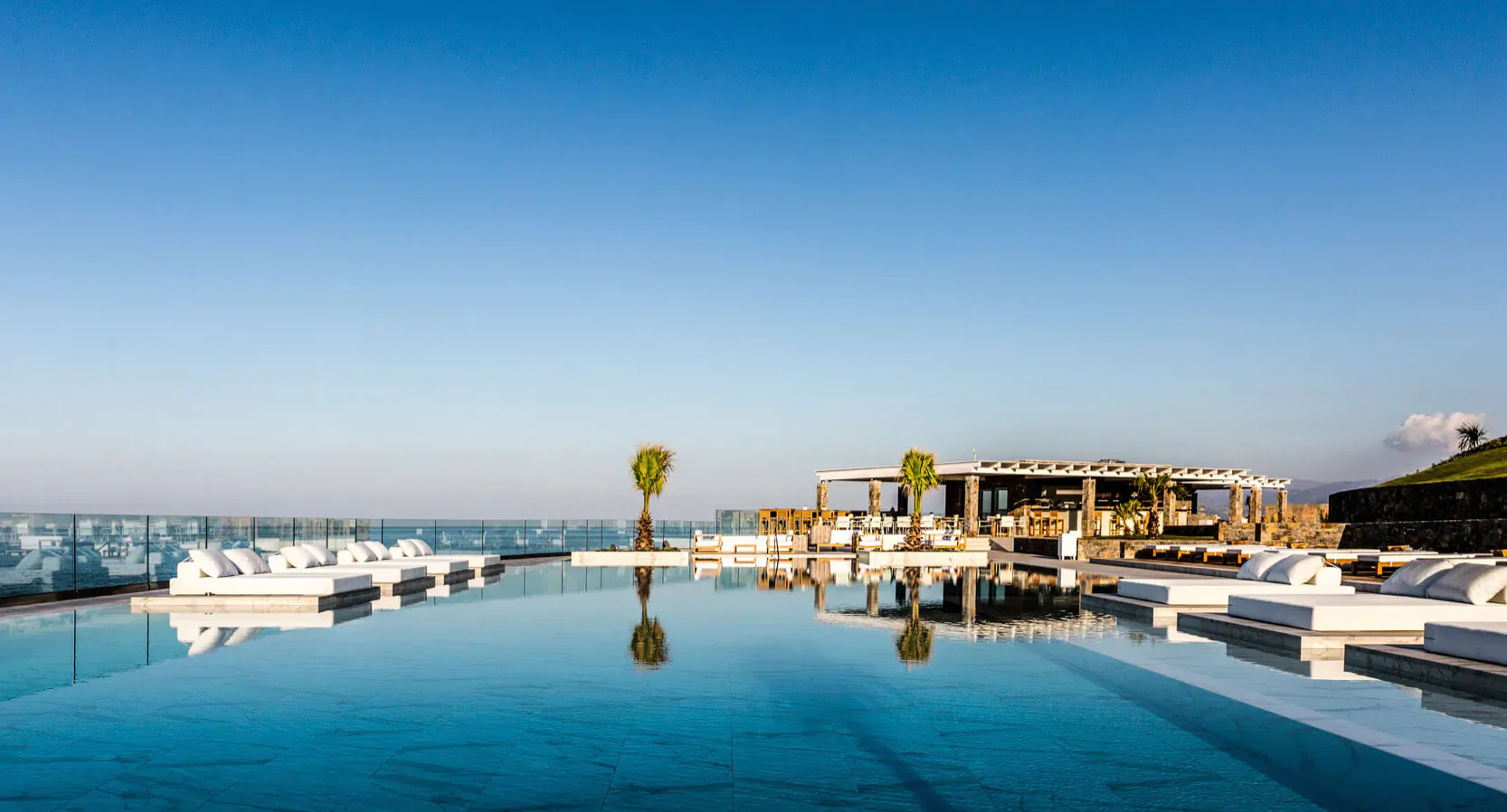 Infinity pool at Abaton Island Resort & Spa with white loungers, palms, pergola, and blue sea view under clear sky.