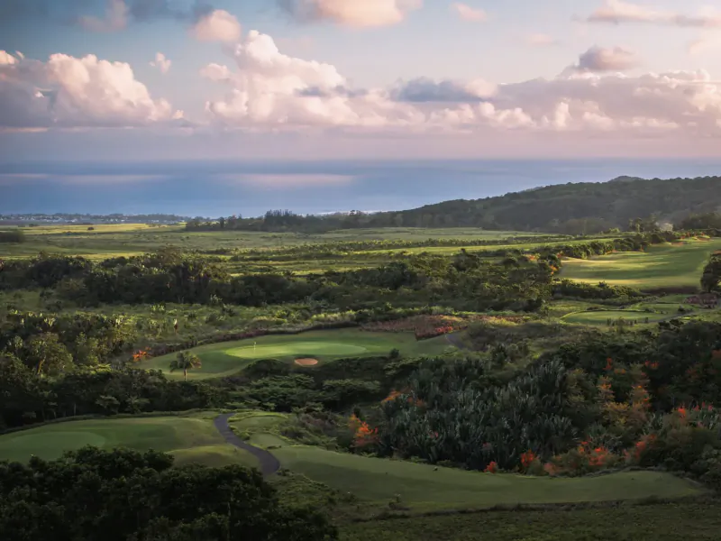 Aerial view of lush green golf course at Avalon Golf Estate, Mauritius, with rolling hills, forests, and sunset sky.