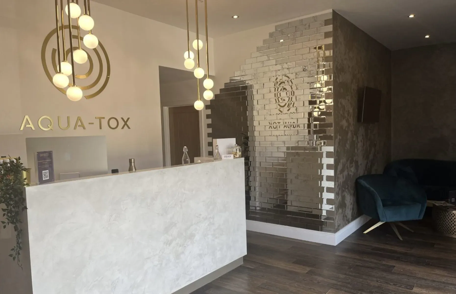 Aqua-Tox Wellness Clinic reception with white marble desk, gold pendant lights, mirrored wall, and green velvet seating.