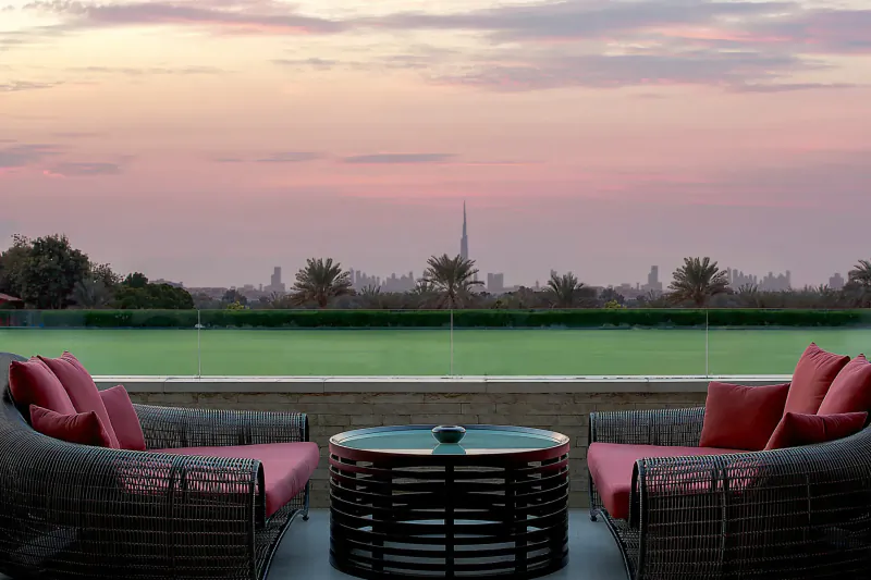 Two red cushioned wicker chairs and low table on terrace overlooking green lawn, palm trees, and Burj Khalifa at sunset, Melia Desert Palm Dubai.