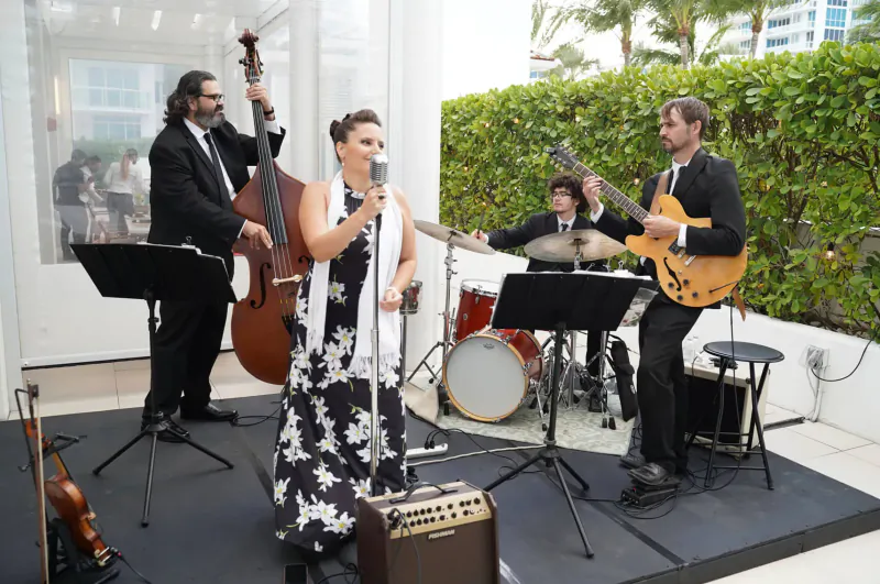 Band performing outdoors at South Beach event: woman singing with mic, bassist, guitarist, drummer on stage amid palms.