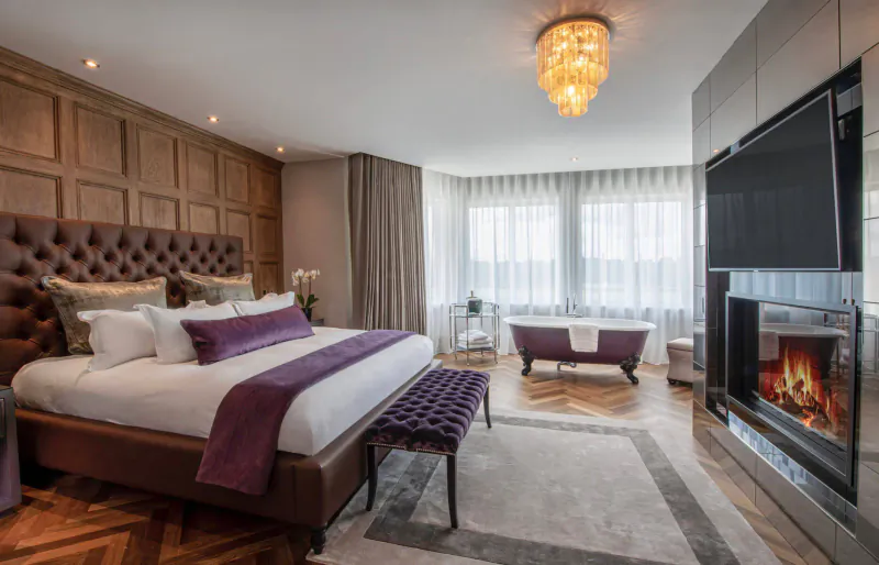 Luxurious hotel bedroom with tufted bed, purple accents, chandelier, freestanding tub, and fireplace.