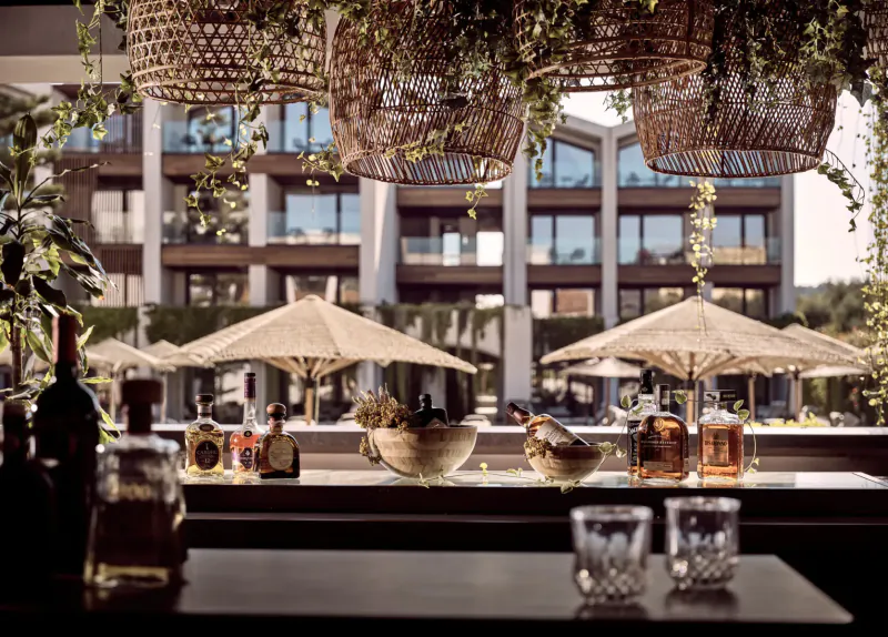 Luxury hotel bar with whiskey bottles, glasses on counter, hanging plants, umbrellas, and modern building backdrop