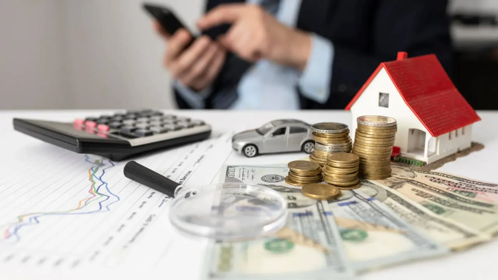 Businessman checking phone near calculator, toy house, car, coins, cash, and magnifying glass on financial chart