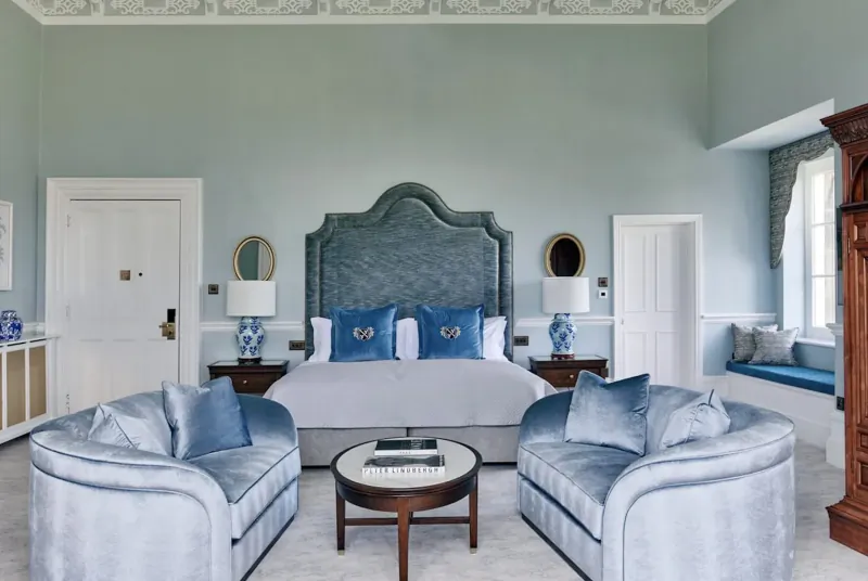 Elegant light blue bedroom in British luxury hotel with tufted king bed, velvet armchairs, round table, lamps, and antique cabinet.