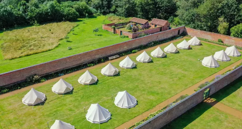 Aerial view of rows of white bell tents in lush green garden enclosed by red brick walls near a house.