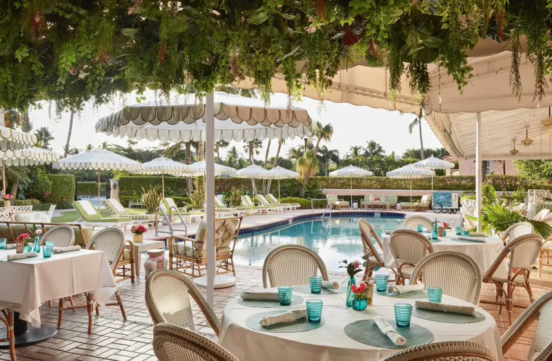Elegant outdoor poolside dining area at Palm Beaches resort with white umbrellas, lush palms, and set tables under draped greenery.