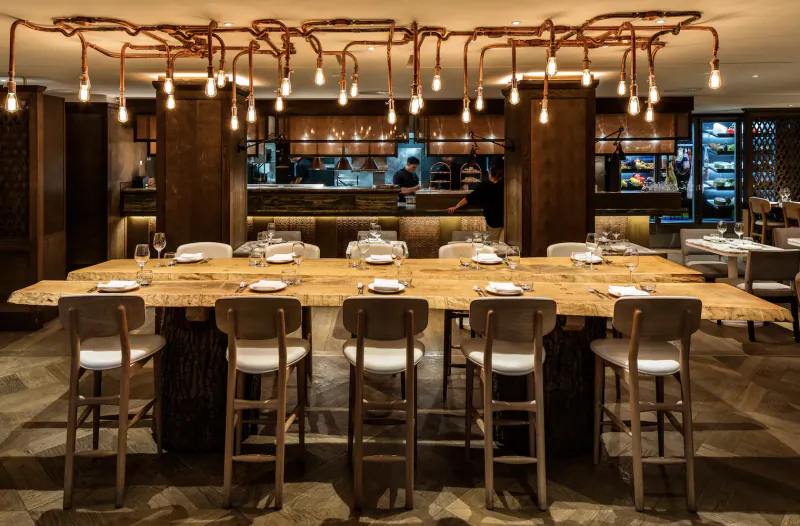 Elegant hotel dining area with large wooden table, high stools, pendant lights, and open kitchen with chefs.