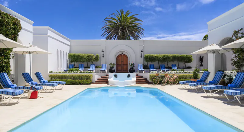 Luxurious courtyard pool at The Marine Hotel Hermanus, with blue loungers, palm tree, white arches, and wooden door.