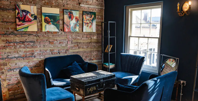 Cozy lounge with navy velvet chairs around vintage trunk table, brick walls, colorful art, shelves, window, lamp.
