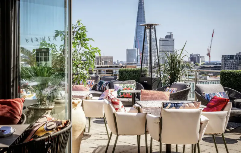 Rooftop terrace with white chairs, colorful cushions, plants, and London skyline including Shard on sunny day