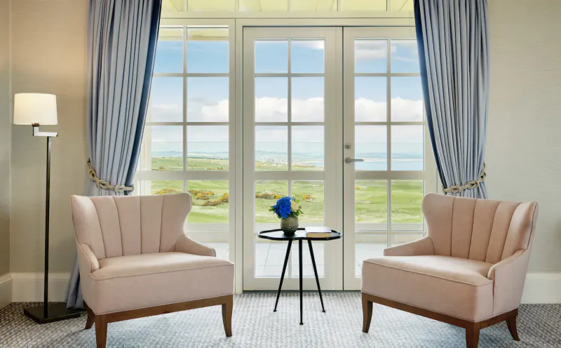 Elegant hotel sitting area with two pink wingback chairs, small table with blue flowers, and French doors open to Scottish golf course view.