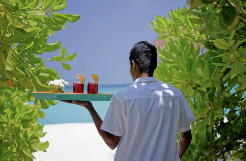 Server in white shirt carrying tray with two red drinks garnished with oranges, tropical beach resort backdrop