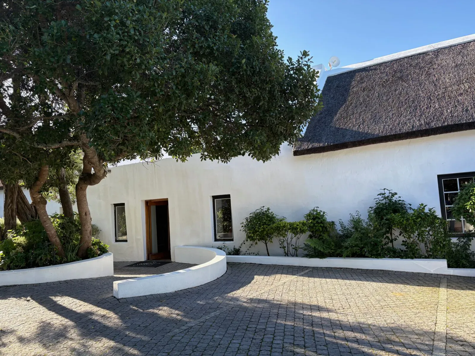 White thatched-roof Old Rectory in South Africa with curved driveway, trees, shrubs, and open wooden door under blue sky.
