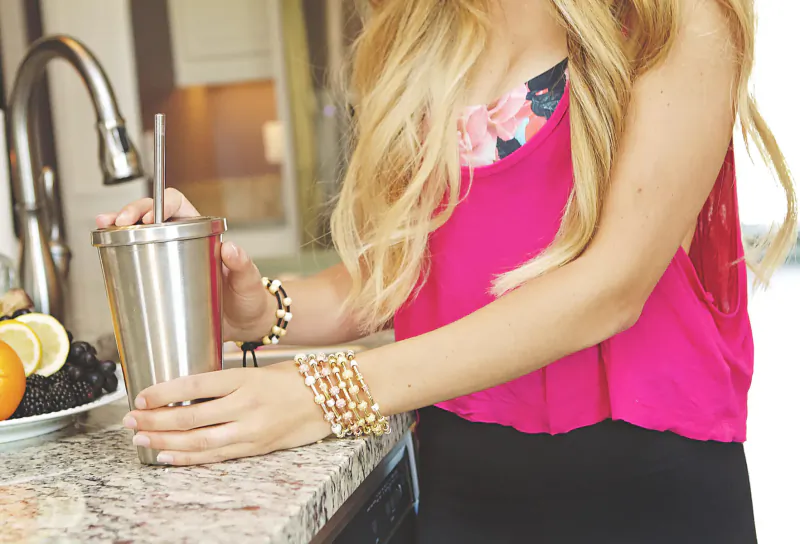 Blonde woman in pink floral tank top holding stainless steel tumbler with straw at kitchen counter, wearing pearl bracelets, lemons nearby