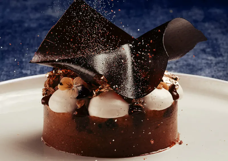 Elegant chocolate entremet on white plate with cream dollops, nuts, viola flowers, and gold dust on blue backdrop