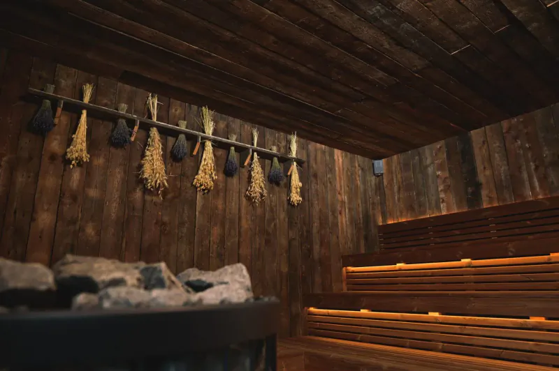 Cozy wooden sauna interior with tiered benches, glowing heater stones, and hanging dried herbs on walls.
