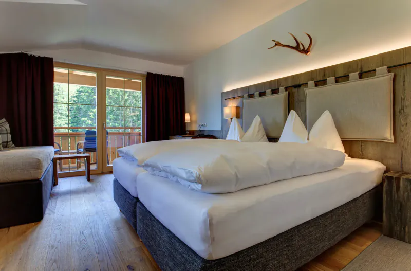 Cozy Hotel Kösslerhof suite with twin beds, antler decor, balcony view of forest and mountains.