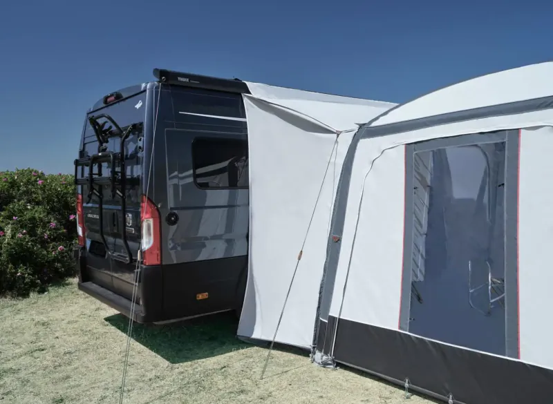 Black motorhome with white Isabella awning extended, bike rack on rear, green bushes, blue sky.