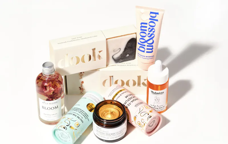 Assortment of Cooks Bloom skincare products: Bloom serum bottle with roses, moisturizers, oils, and creams on white surface.