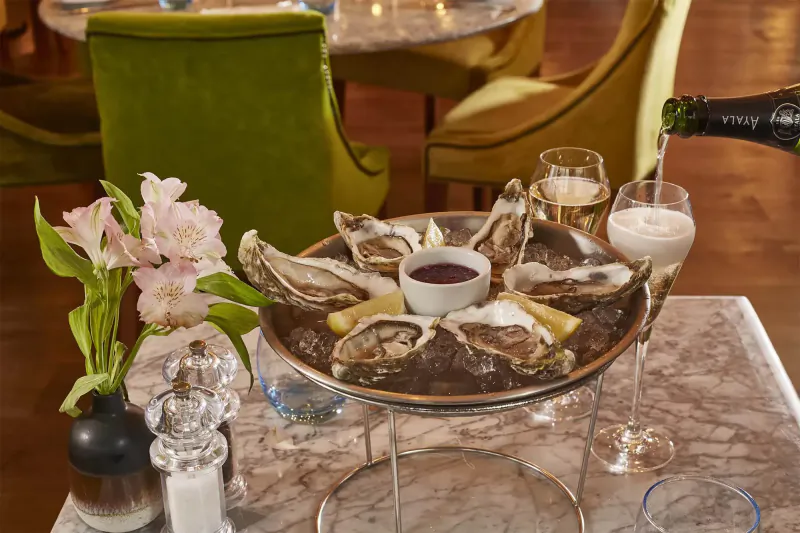 Oysters on ice with lemon and sauce in gold stand, champagne poured into glasses, pink flowers, at elegant restaurant table