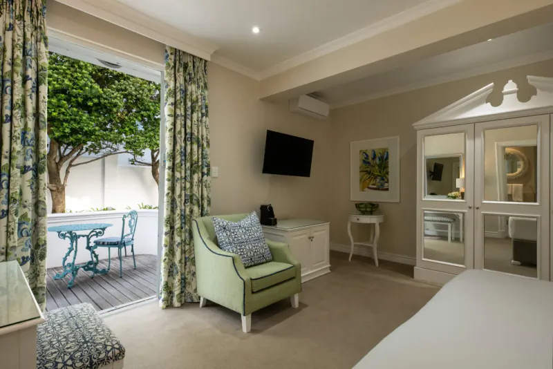 Elegant hotel room at The Marine with open doors to leafy balcony, green armchair, bed, and TV.