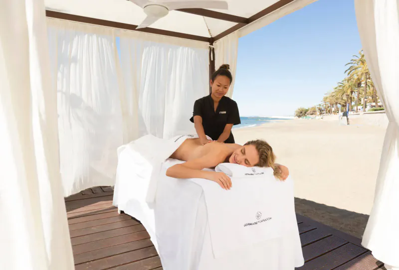 Asian masseuse giving shoulder massage to blonde woman on beachside spa bed under white cabana, Amàre logo, ocean view.