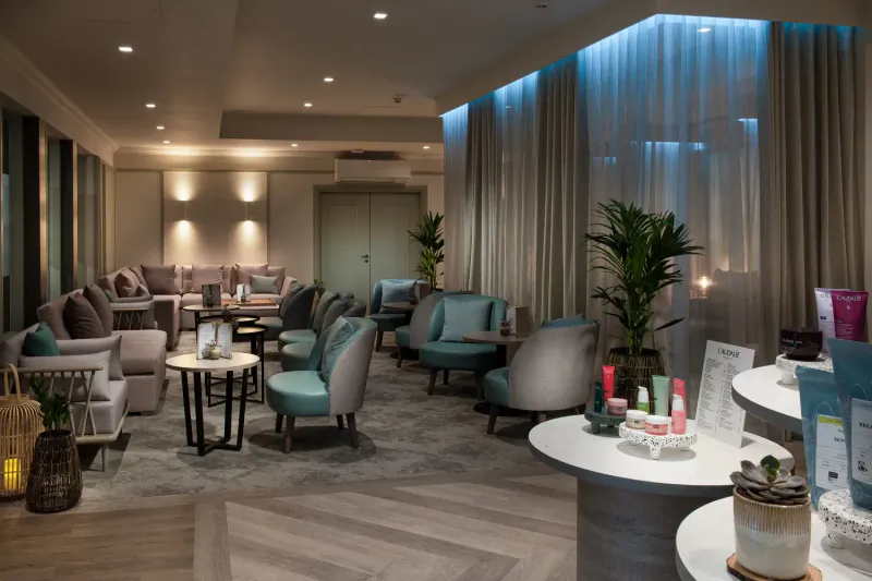 Elegant hotel spa lounge with plush blue-grey armchairs, plants, candles, skincare products, and ambient blue lighting.