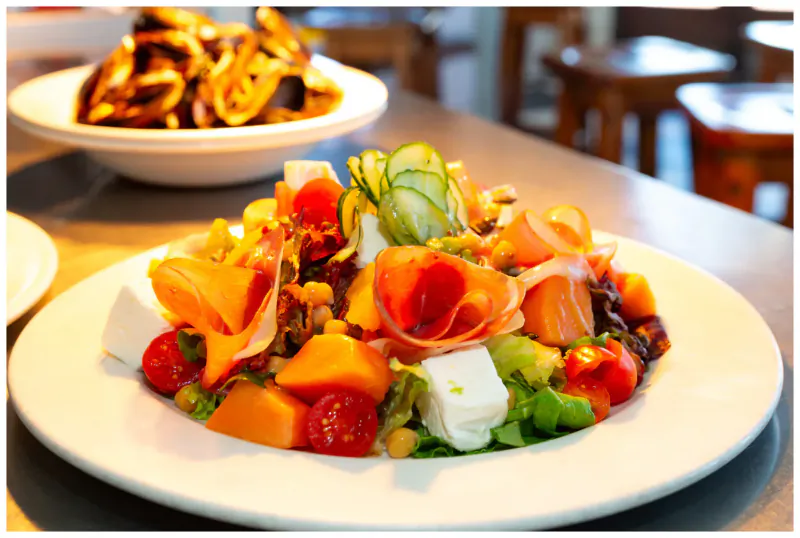 Vibrant salmon salad with feta, tomatoes, zucchini, nuts on white plate, side bowl of fried onions at café table.