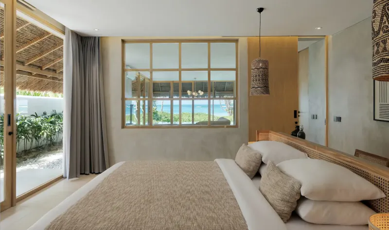 Spacious beachfront bedroom at Amber Lombok Resort with king bed, large window to ocean, thatched roof, and tropical decor.