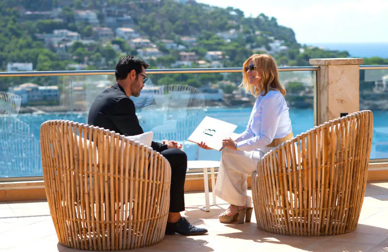 Man in suit and woman in white blouse discuss holding papers on terrace overlooking sea and hills