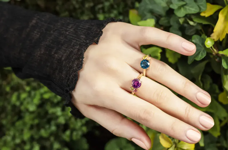 Woman's hand wearing two gold rings with teal and purple gemstones against green foliage