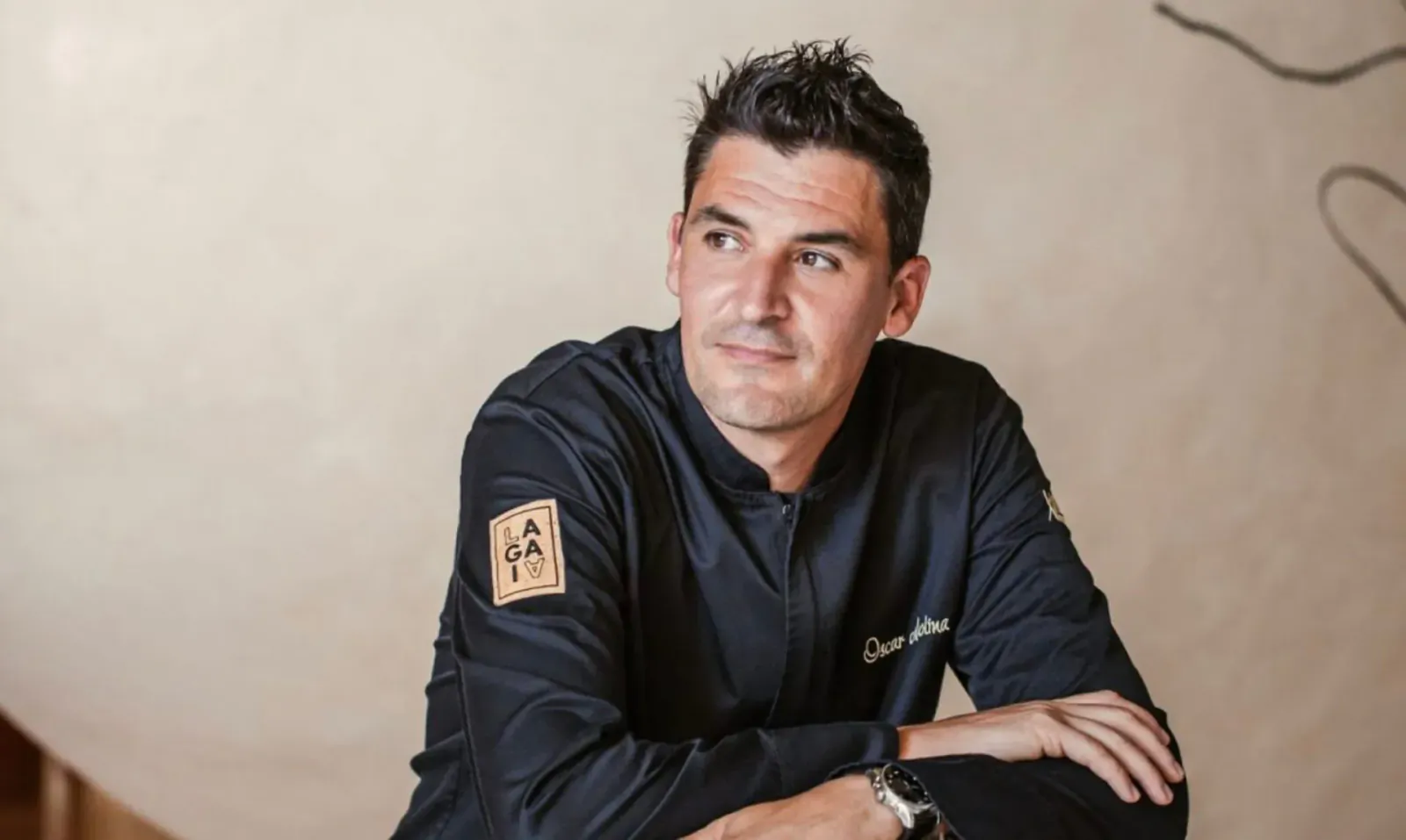 Smiling chef in black uniform with 'Ibiza Gran Hotel' embroidered, arms crossed, looking aside in elegant setting.