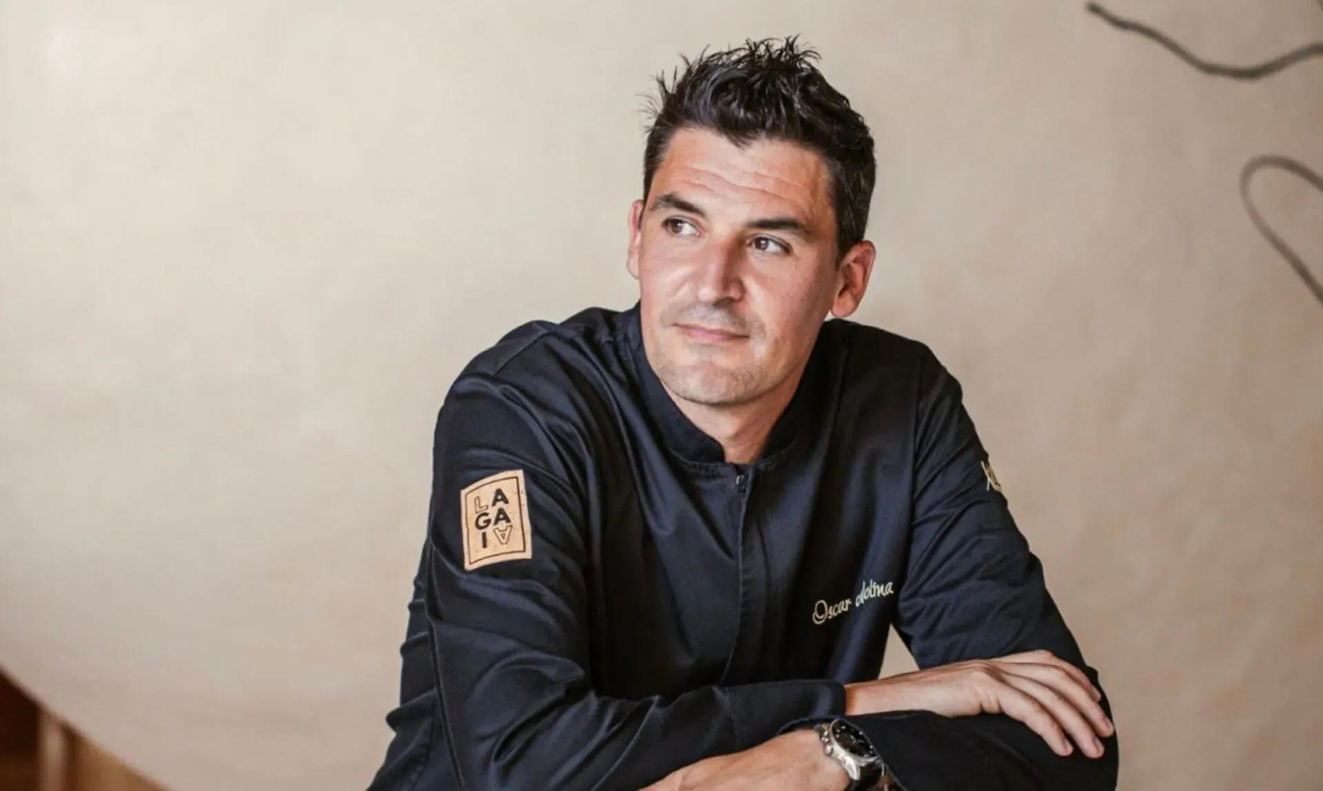 Smiling chef in black uniform with 'Ibiza Gran Hotel' embroidered, arms crossed, looking aside in elegant setting.